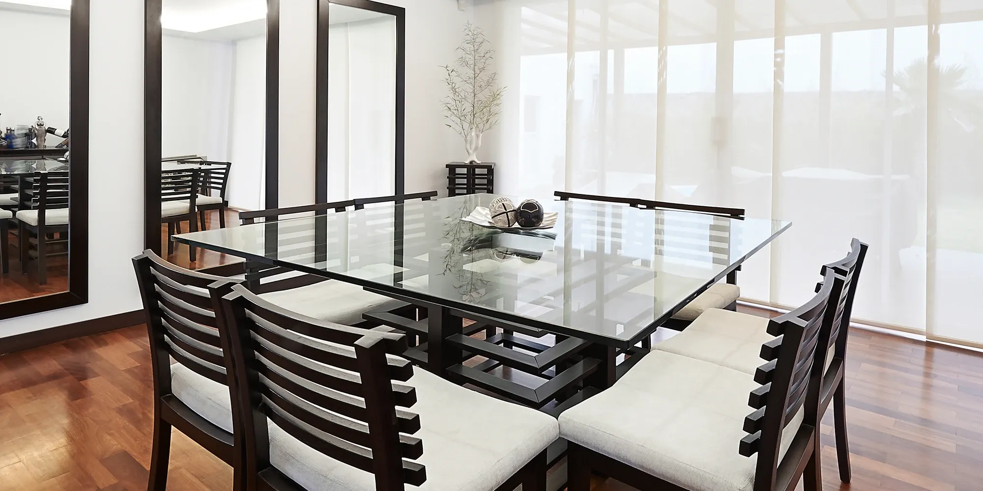 A sophisticated dining room featuring a large, rectangular glass table top on a dark wood base, surrounded by matching chairs on a hardwood floor.