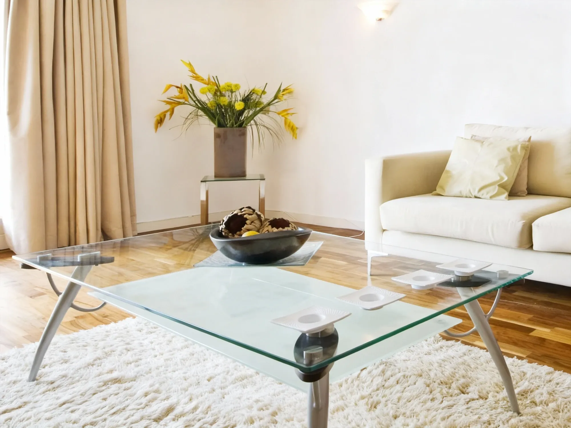 A modern living room with a two-tiered glass coffee table, featuring a clear top and a frosted lower shelf, set on a white shag rug.