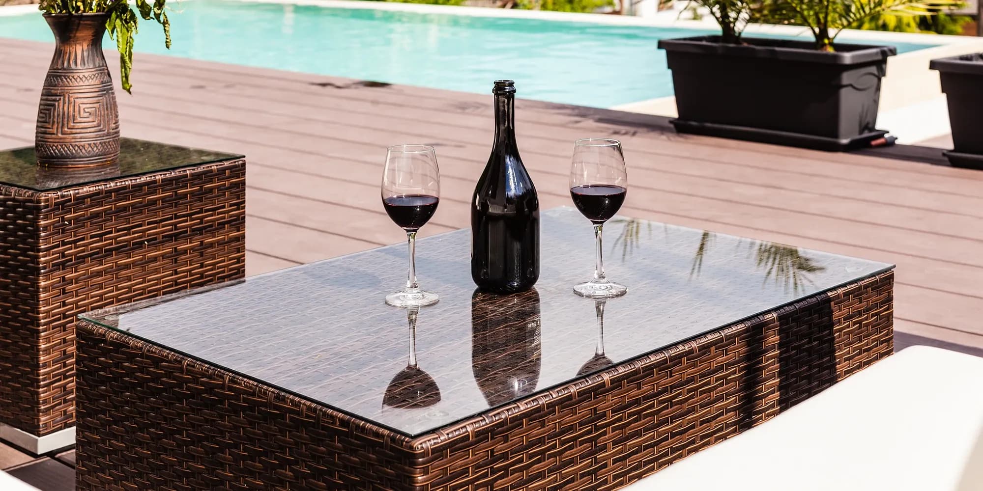 A close-up of a glass-top wicker coffee table on a poolside deck, set with a bottle of red wine and two glasses, ready for relaxation.