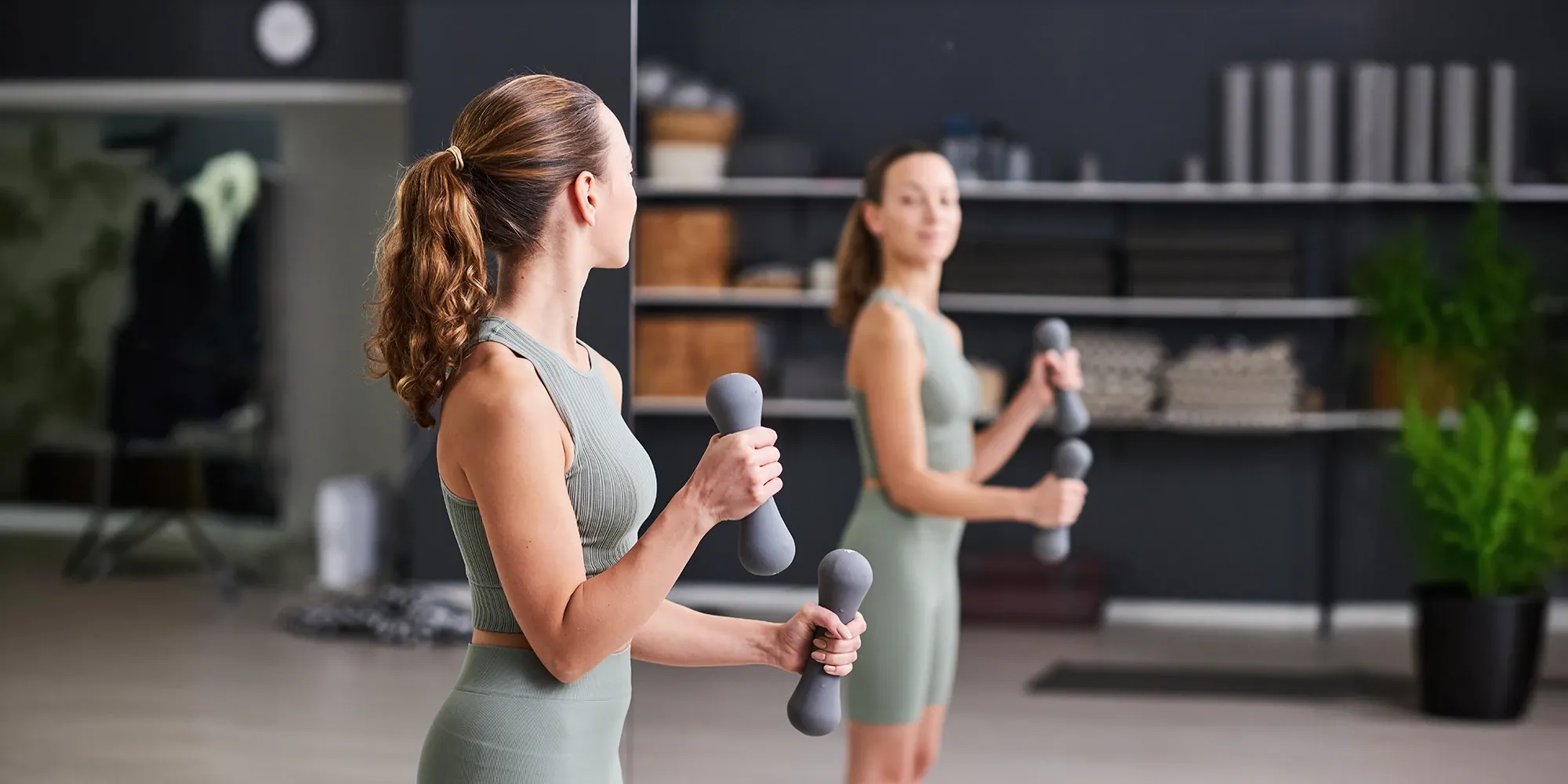 A woman in activewear lifts dumbbells while watching her form in a large, clear gym mirror, which reflects her focused expression.