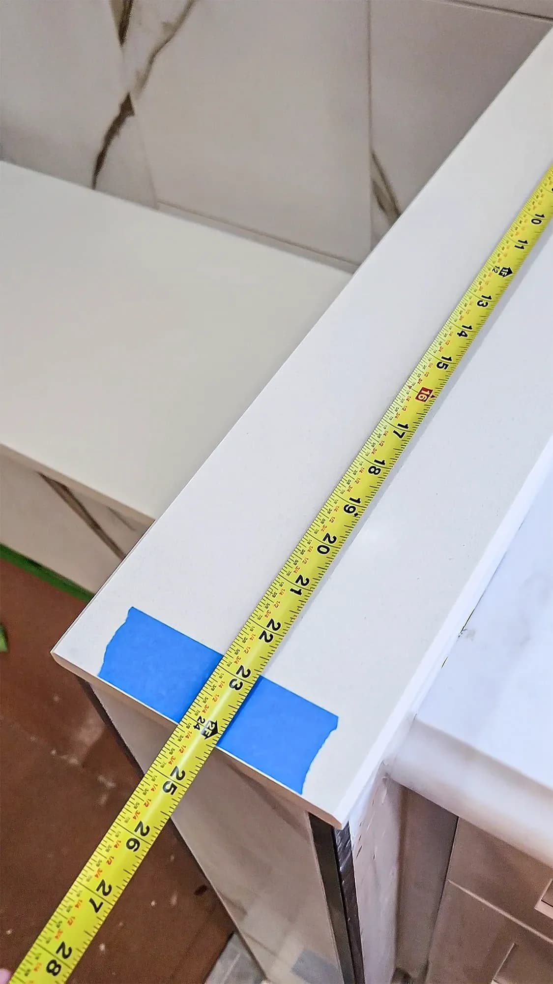 Close-up of a yellow tape measure checking the length of a white bathroom half-wall, showing a measurement of approximately 24 inches.