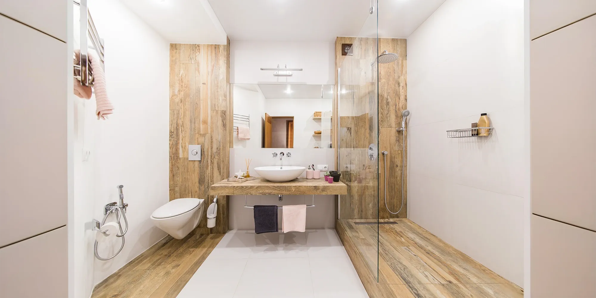 A spa-like modern bathroom with a walk-in shower separated by a frameless glass panel, featuring extensive use of light wood-look tiles and a floating vanity with a vessel sink.