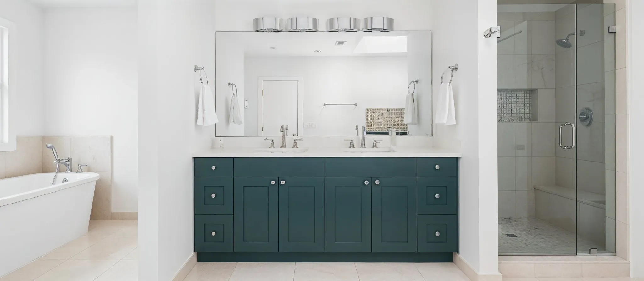 A bright and modern bathroom featuring a stylish dark teal double vanity, a large frameless wall mirror, and a walk-in glass shower.