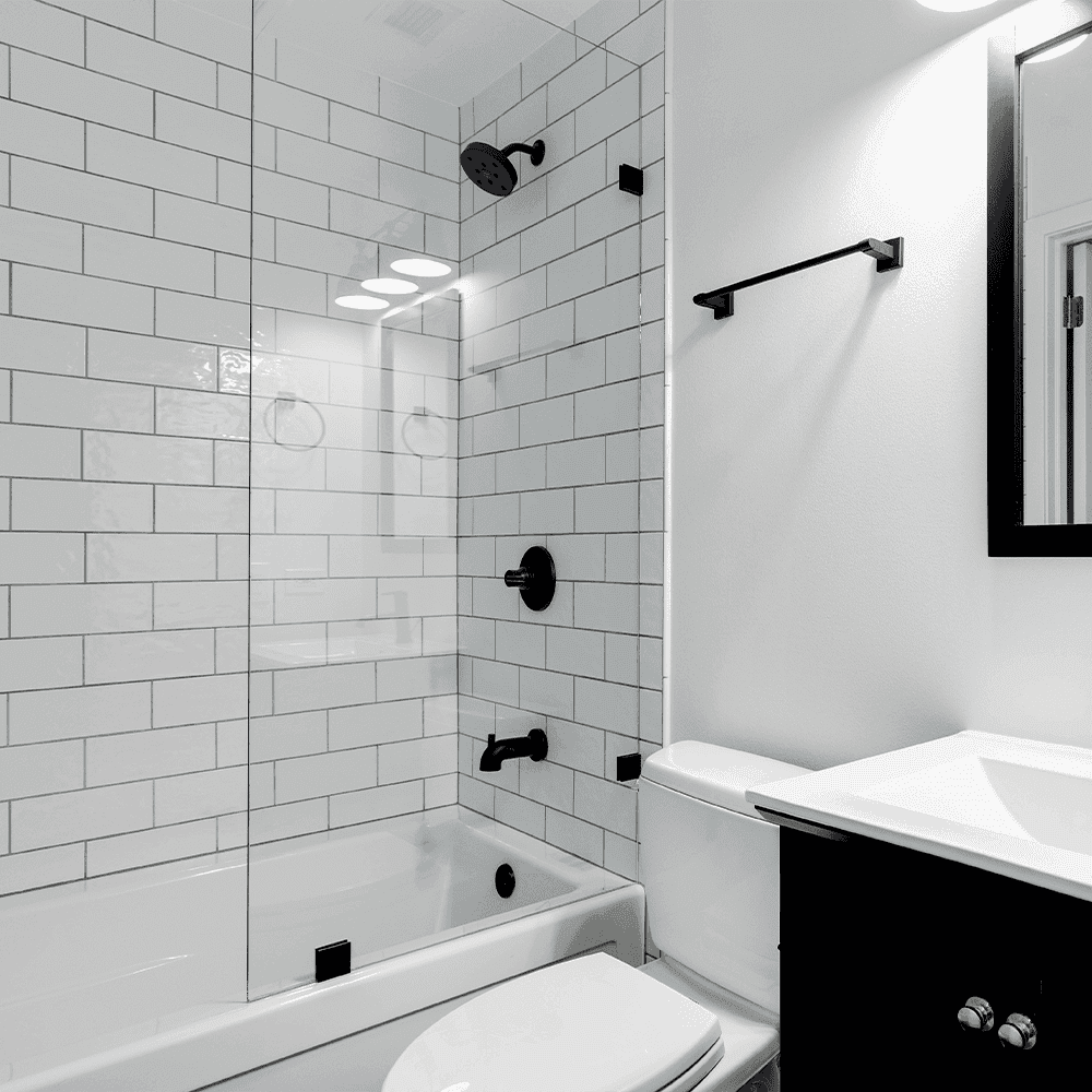 Clear tempered glass shower door in modern bathroom with white subway tile, black fixtures, and beveled edge mirror, showcasing custom glass solutions.