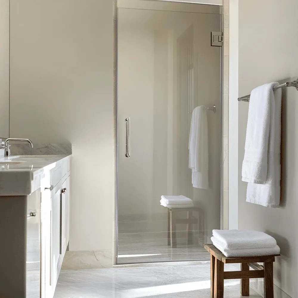 Minimalist bathroom with a custom glass shower, marble floor, and neatly stacked white towels on a wooden stool.