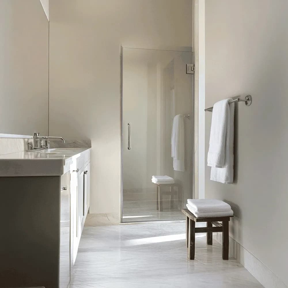 Modern bathroom with a custom glass shower enclosure, marble floor, and neatly folded white towels on a wooden stool.