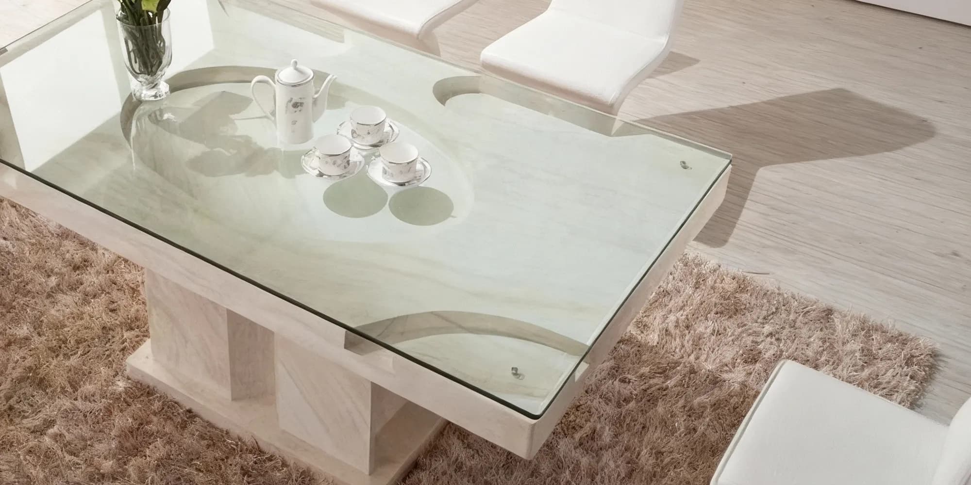A modern dining table with a thick, clear glass table cover protecting a light-colored stone base, set with a white teapot and cups.