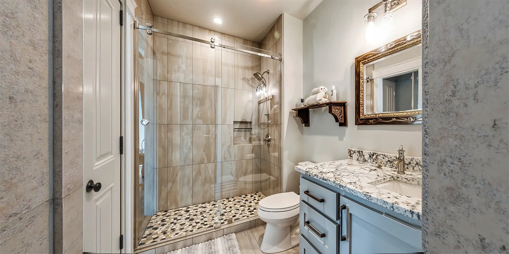 A modern bathroom featuring a walk-in shower with a sliding glass door, wood-look wall tiles, and a river rock pebble floor, next to a grey vanity with a quartz countertop.