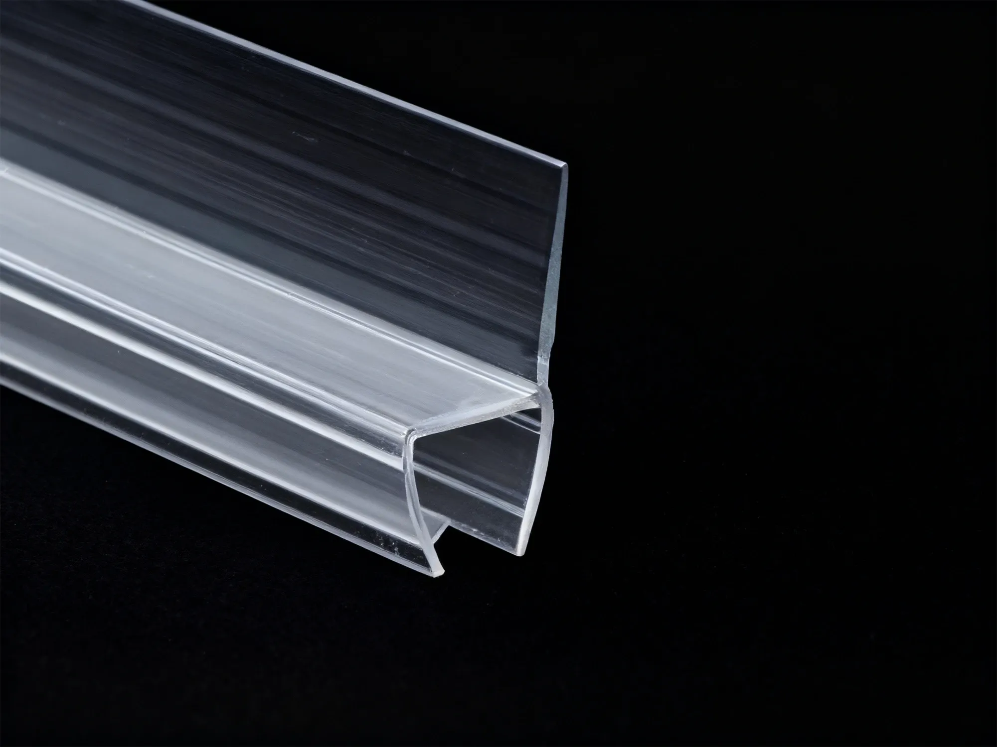 A close-up macro shot of a clear, pre-formed vinyl shower door sweep with a drip rail, shown against a solid black background.