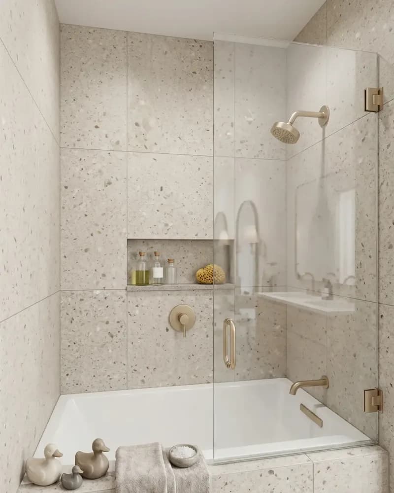 Fixed glass shower panel over a bathtub with beige terrazzo tile and brass hardware