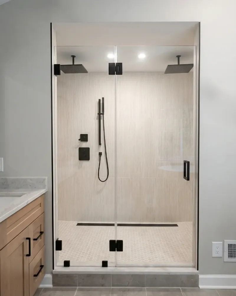 Frameless hinged glass shower door with dual shower heads and matte black hardware on wood-look tile