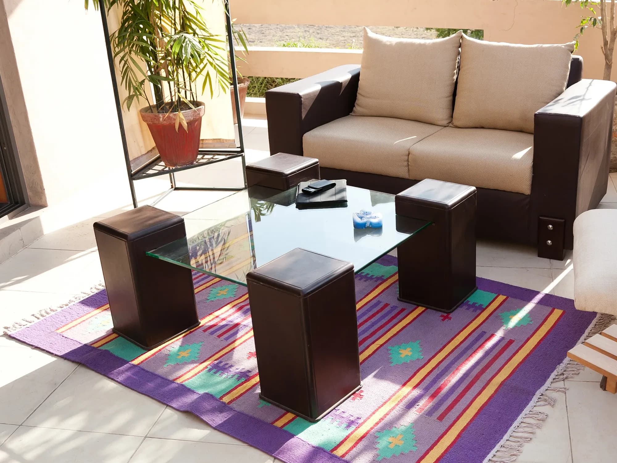 A sunny patio featuring a modern sofa and a square glass-top coffee table with four matching ottomans arranged on a colorful striped rug.