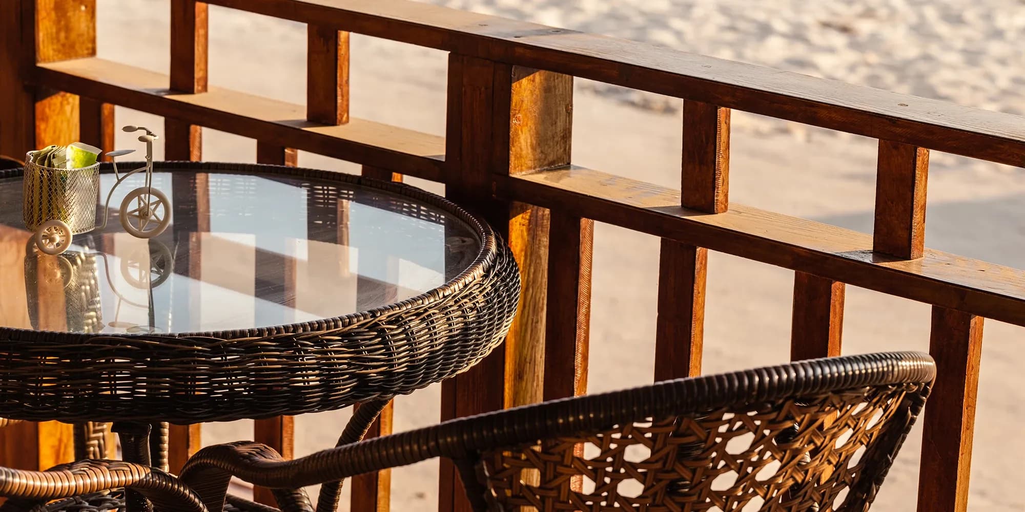 A close-up of a dark wicker patio table with a clear, reflective glass top, set on a wooden balcony overlooking a sunny beach.