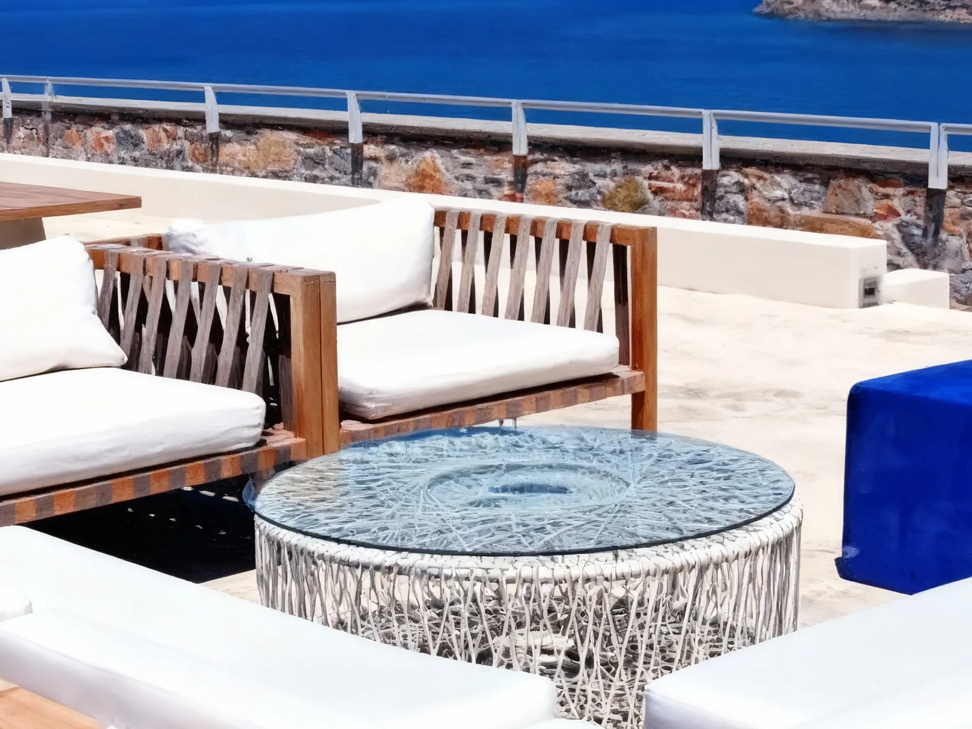 A modern white woven patio coffee table with a round glass top, part of a lounge set overlooking a deep blue sea.