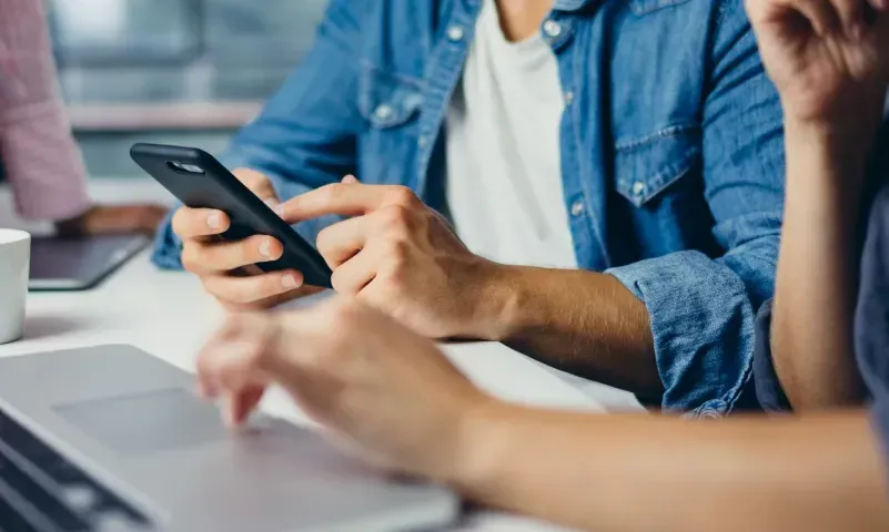 Two people engaged in conversation while using a smartphone and a laptop, representing collaboration in custom glass solutions, including shower doors, table tops, mirrors, and tempered glass products.