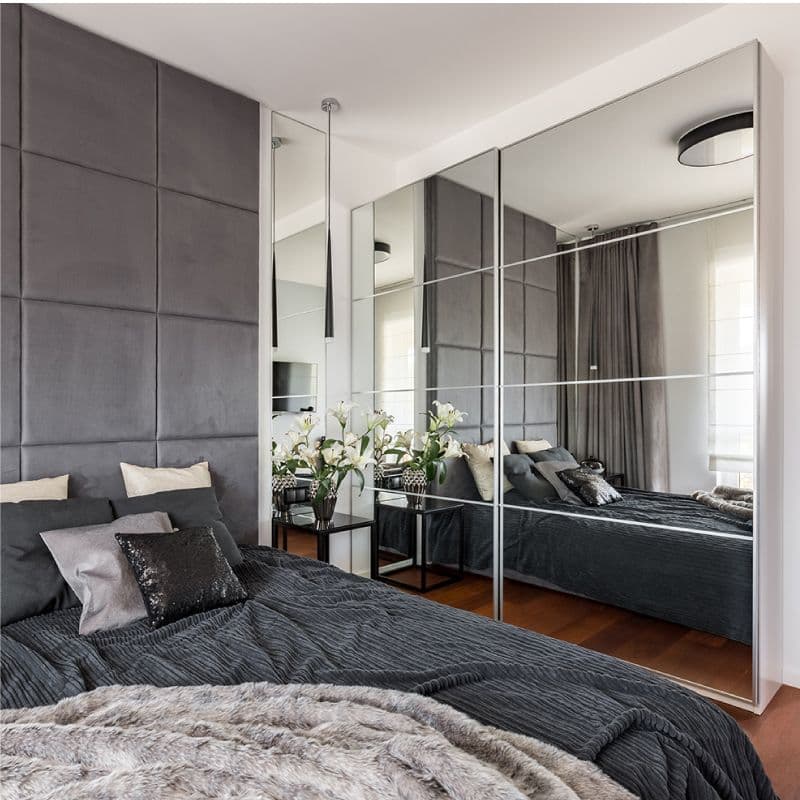 Modern bedroom with custom mirrors reflecting elegant decor, featuring a stylish bed, a black nightstand with flowers, and textured bedding, highlighting sophistication with tempered glass accents.