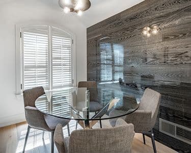 A modern dining area featuring a round glass table with a beveled glass edge, surrounded by stylish chairs, and illuminated by contemporary lighting against a dark wood accent wall, highlighting the elegance of custom glass design.