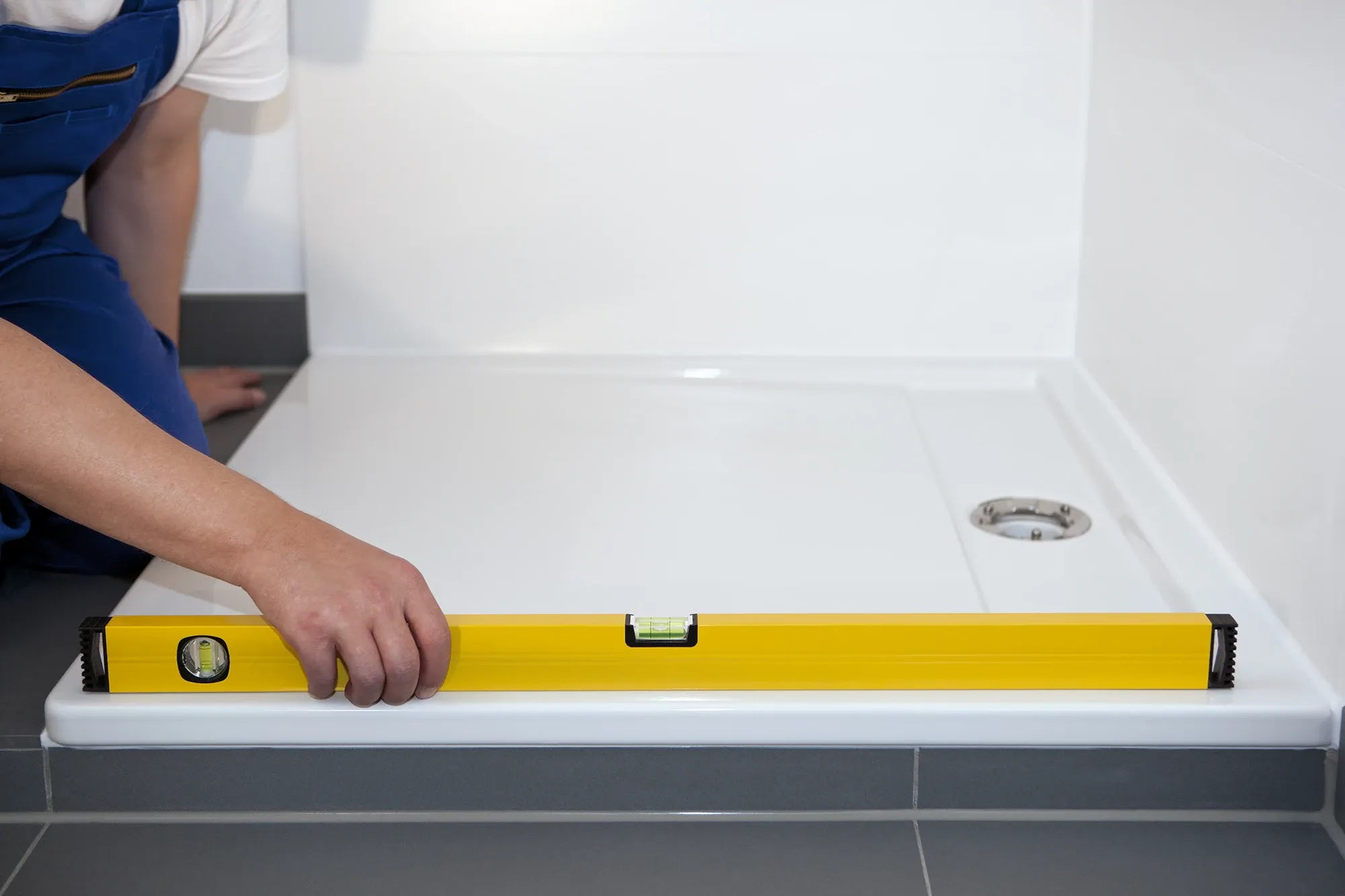 A plumber uses a yellow bubble level to ensure a white shower tray is installed correctly on a dark tiled floor.