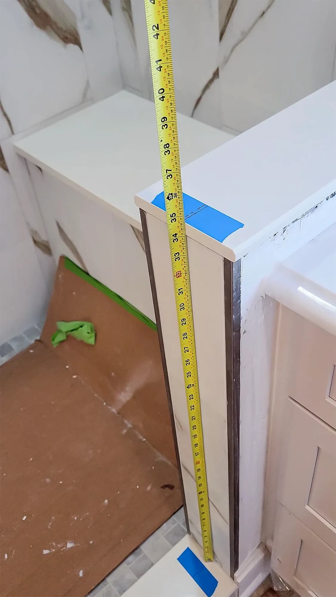 A yellow tape measure is used to check the height of a white bathroom pony wall for a custom shower glass installation.