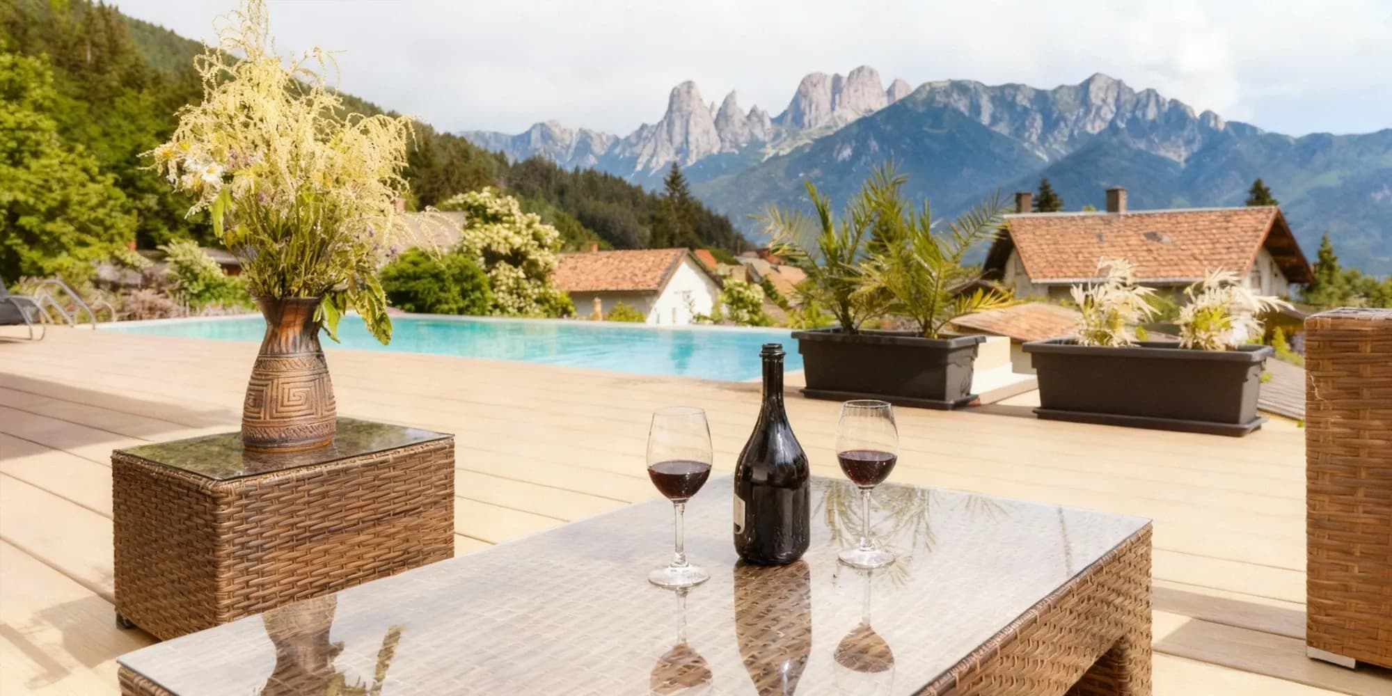 A glass-top wicker coffee table set with a wine bottle and two glasses, on a poolside deck with a breathtaking mountain view.